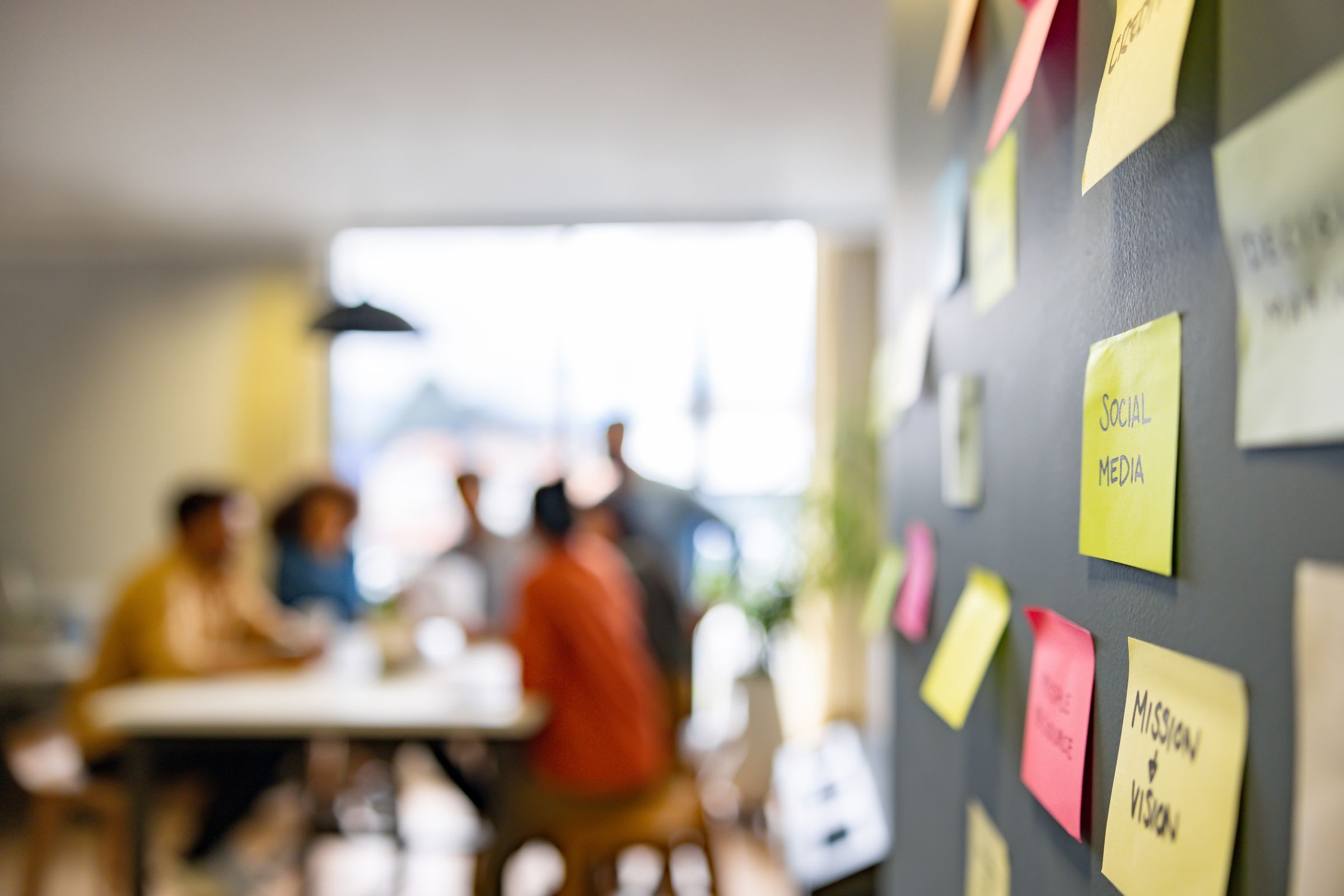 Sticky notes on a wall in sharp focus, with a team meeting blurred in the background